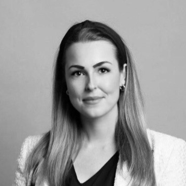 Schwarz-Weiß-Porträt einer Frau mit langem glattem Haar, die einen hellen Blazer über einem dunklen Oberteil trägt und mit einem leichten Lächeln in die Kamera blickt, vor einem schlichten Hintergrund.