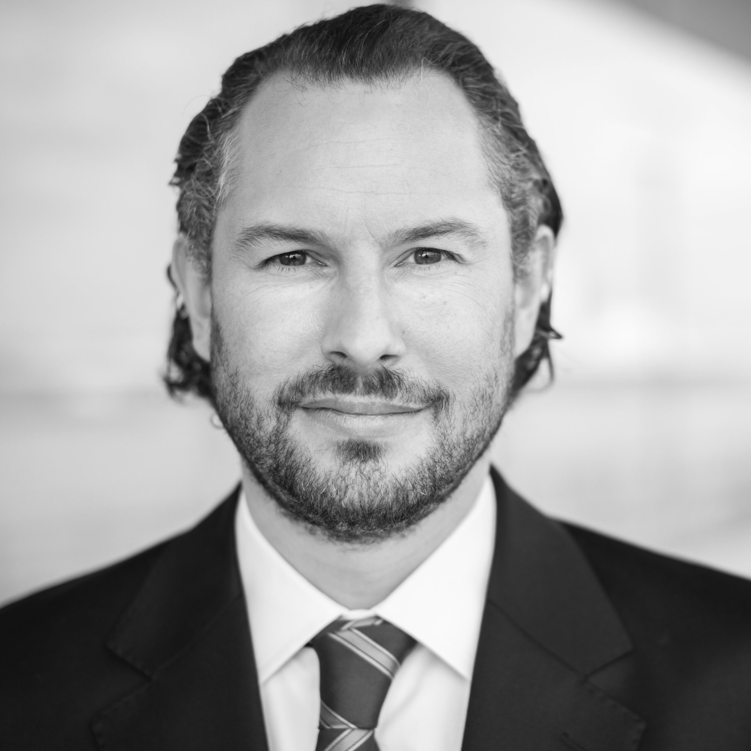 Ein Mann mit gewelltem dunklem Haar und Bart, der einen Anzug und eine gestreifte Krawatte trägt, lächelt leicht in die Kamera. Der Hintergrund ist unscharf und in hellen Farben gehalten. Das Bild ist in schwarz-weiß gehalten.