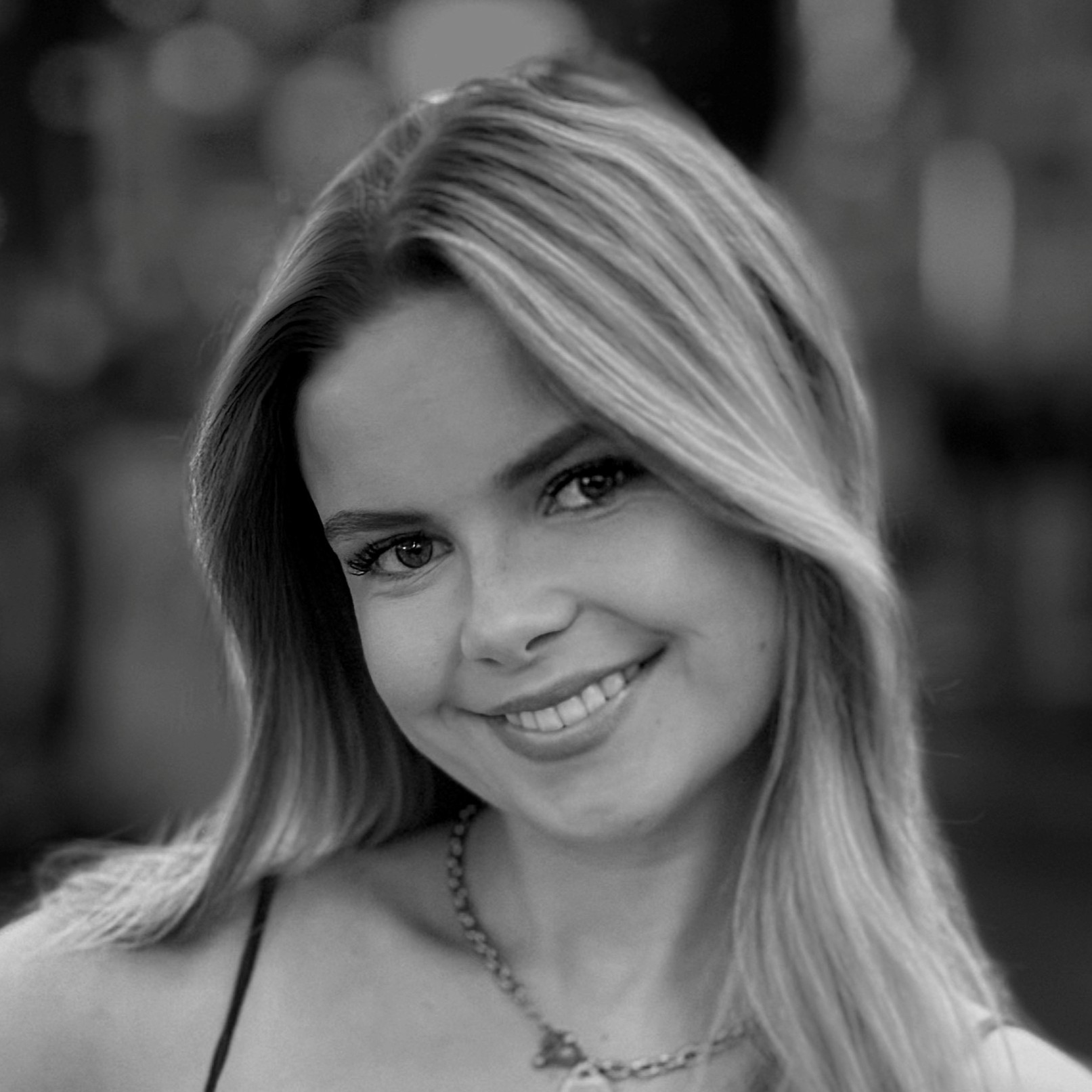 Eine junge Frau mit langem, glattem Haar lächelt in die Kamera. Sie trägt eine Halskette und ein Oberteil mit dünnen Trägern. Der Hintergrund ist unscharf, um die Aufmerksamkeit auf ihr Gesicht zu lenken. Das Bild ist in schwarz-weiß gehalten.