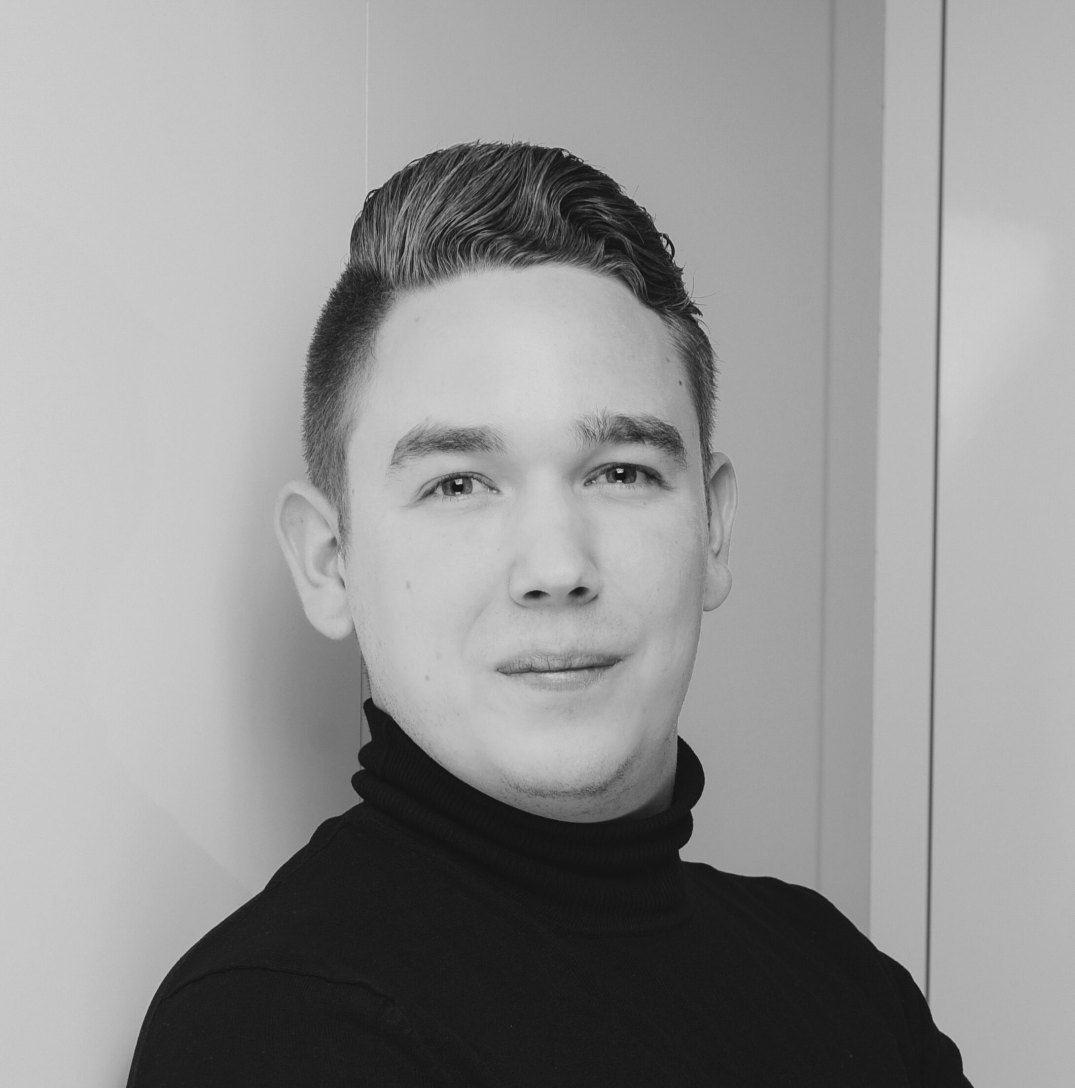 Ein junger Mann mit kurzem, gestyltem Haar trägt einen schwarzen Rollkragenpullover und blickt mit einem leichten Lächeln in die Kamera. Der Hintergrund ist schlicht und minimalistisch. Das Foto ist in schwarz-weiß gehalten.