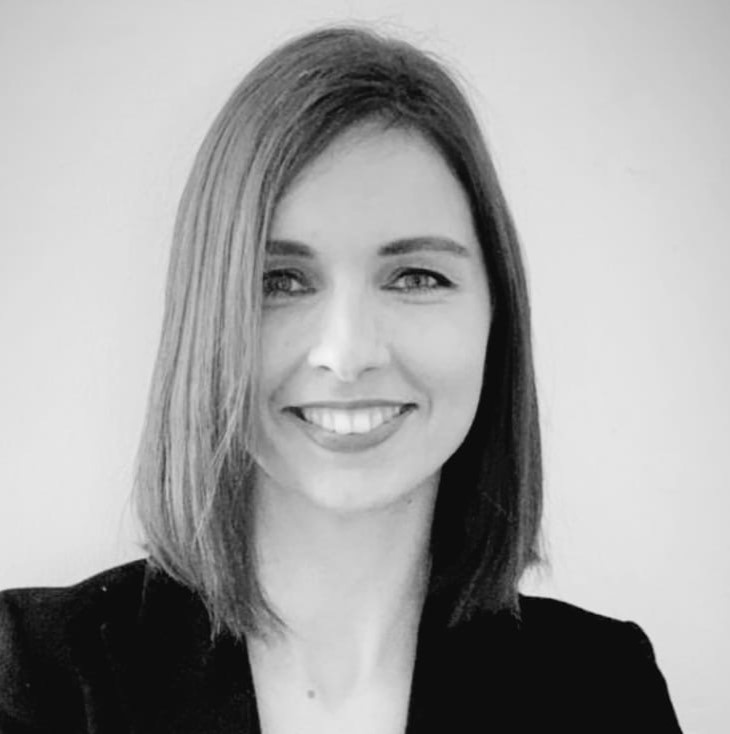 Schwarz-Weiß-Porträt einer Frau mit glattem, schulterlangem Haar, die in die Kamera lächelt und einen dunklen Blazer trägt. Der Hintergrund ist schlicht und hell.