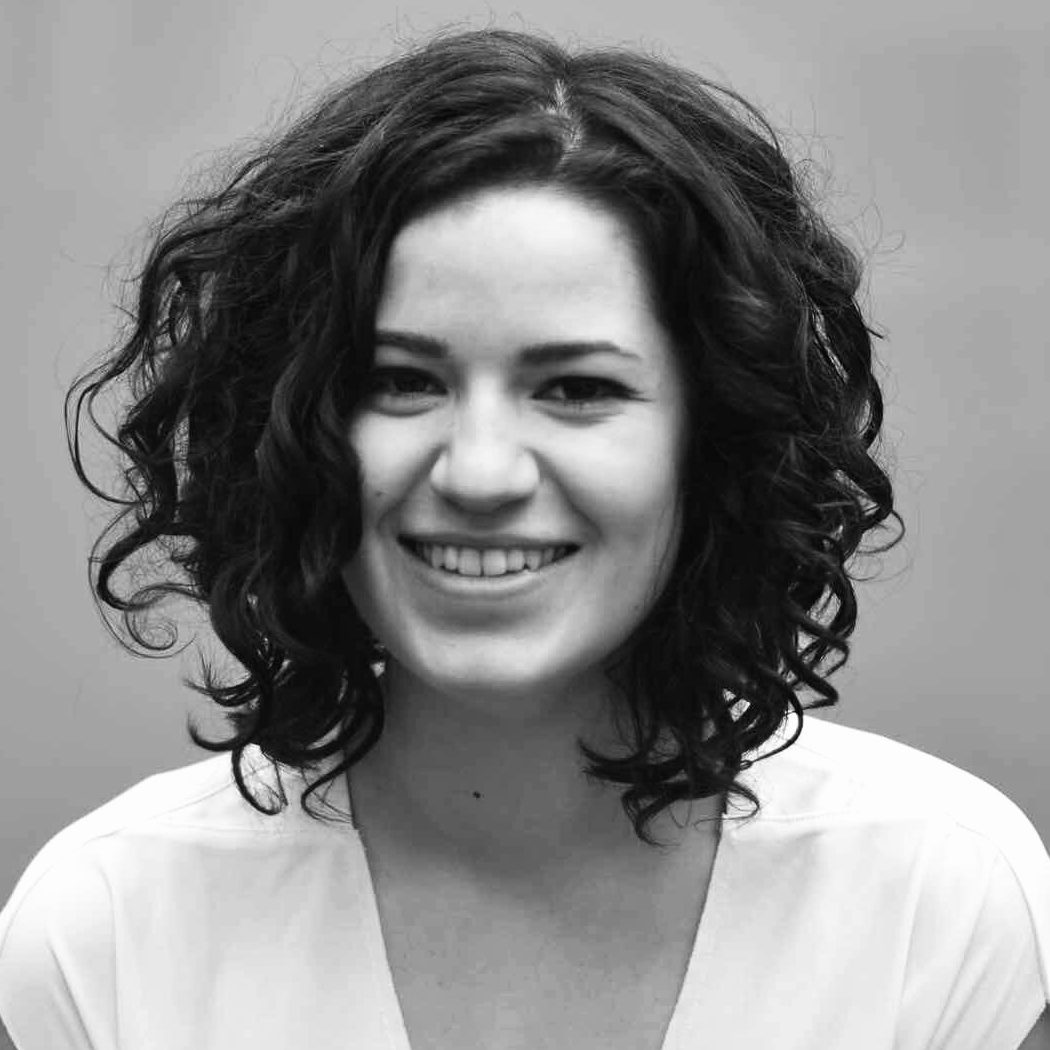 Eine Frau mit lockigem dunklem Haar lächelt in die Kamera. Sie trägt ein helles Oberteil und der Hintergrund ist schlicht und unscharf. Das Bild ist in schwarz-weiß gehalten.