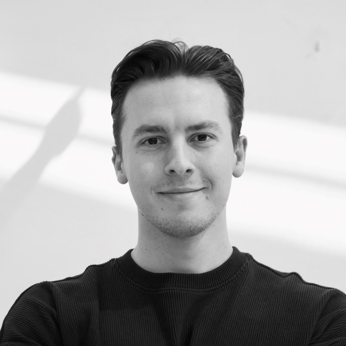 Ein junger Mann mit dunklem Haar, der einen dunklen Pullover trägt, lächelt sanft, während er vor einem schlichten hellen Hintergrund mit diagonalen Schatten steht.