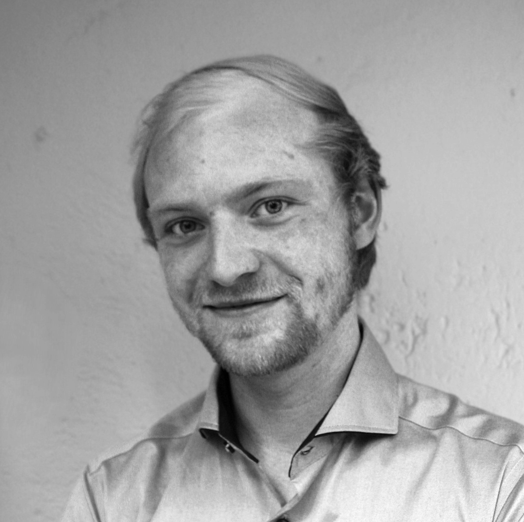 Ein Mann mit hellem Haar und einem kurzen Bart lächelt leicht, trägt ein helles Hemd mit Kragen und posiert vor einer einfarbigen, strukturierten Wand. Das Foto ist in schwarz-weiß gehalten.