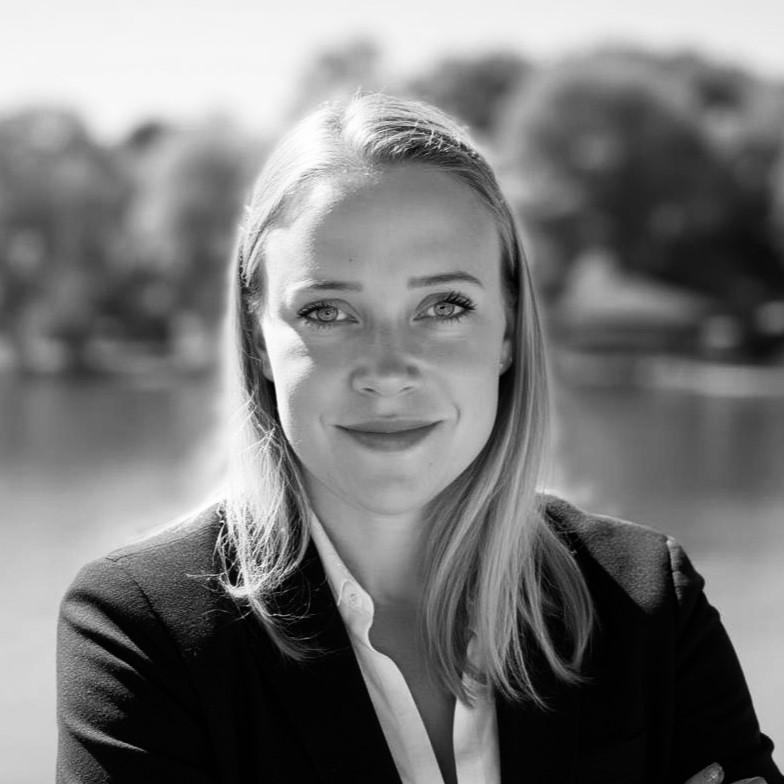 Eine Frau mit glattem, hellem Haar, die einen dunklen Blazer und ein weißes Hemd trägt, lächelt sanft in die Kamera. Sie steht im Freien vor einem unscharfen Hintergrund mit Bäumen und Wasser. Das Foto ist in schwarz-weiß gehalten.