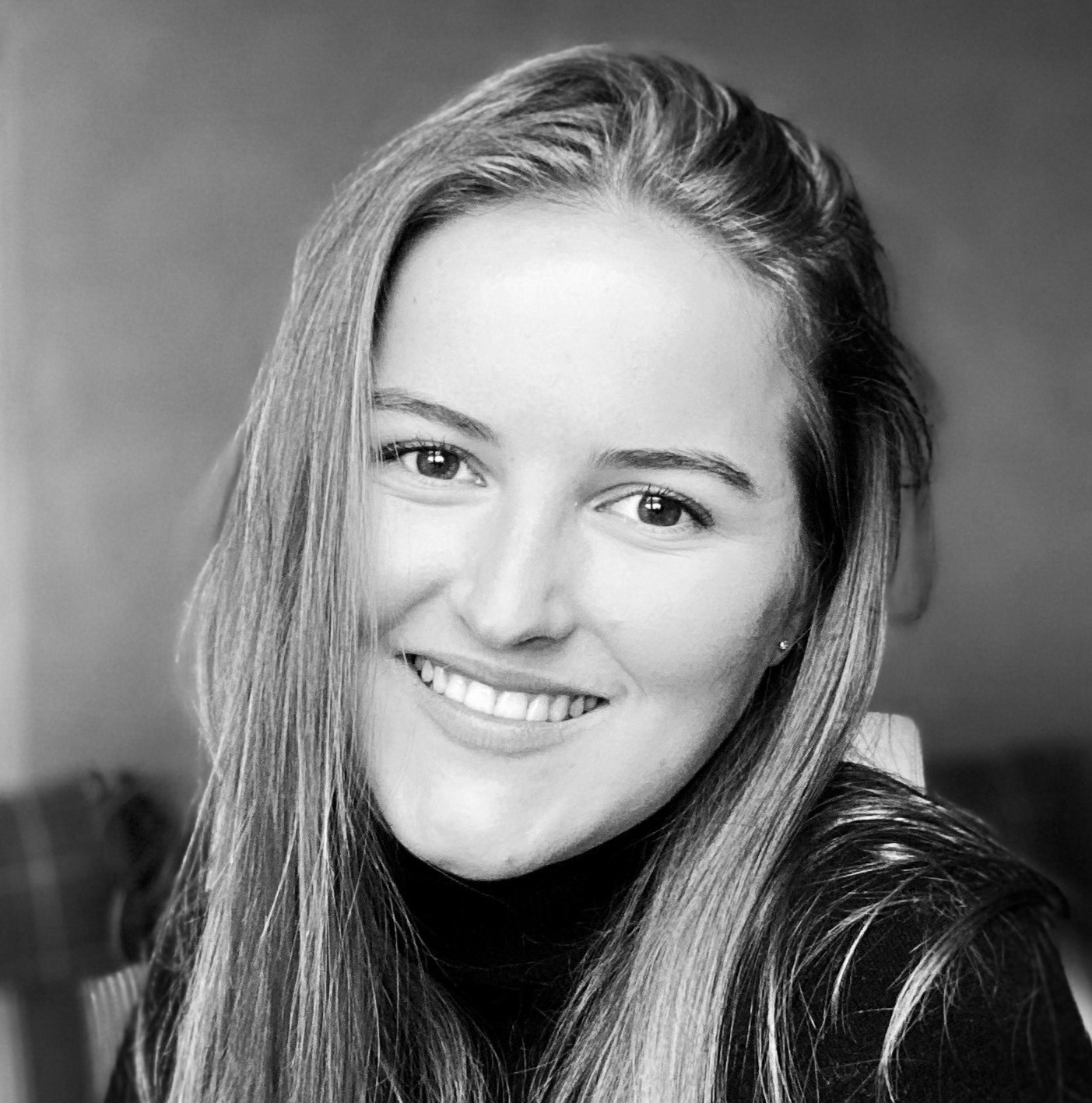 Schwarz-weißes Porträt einer jungen Frau mit langen Haaren, die in die Kamera lächelt. Sie trägt ein dunkles Oberteil, und der Hintergrund ist leicht unscharf.